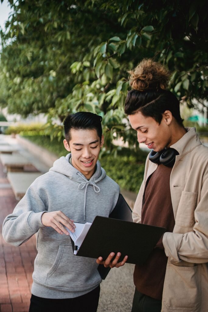 Content diverse male students wearing casual outfits reading new material in folder while standing together in lush green park